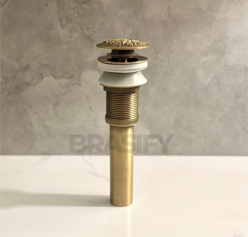 May include: A gold-toned drain stopper with a decorative top and a white ceramic ring. The drain stopper is made of metal and has a cylindrical shape.