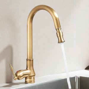 May include: A gold-toned kitchen faucet with a high-arc spout. The faucet has a lever handle and a pull-down sprayer. Water is flowing from the spout into a stainless steel sink. The faucet is mounted on a light-colored wall.