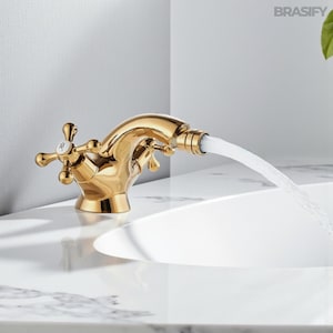 Può includere: Un rubinetto dorato con maniglie a forma di croce e un beccuccio curvo. L'acqua scorre dal beccuccio in un lavandino bianco. Il rubinetto ha una superficie lucida e riflettente. Il marchio "BRASIFY" è visibile in alto a destra.