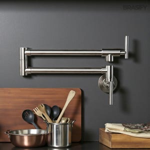 May include: A stainless steel pot filler faucet mounted on a dark gray wall. The faucet has a long, articulated arm and a single handle. Kitchen utensils and a copper-colored bowl are in the foreground.