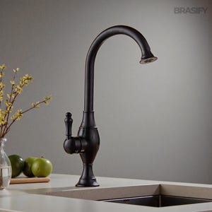 Könnte beinhalten: Ein dunkelbronze Küchenarmatur mit gebogenem Auslauf und dekorativem Griff. Die Armatur ist auf einer hellen Arbeitsplatte montiert, darunter ist ein schwarzes Waschbecken zu sehen. Der Markenname "BRASIFY" befindet sich oben rechts.