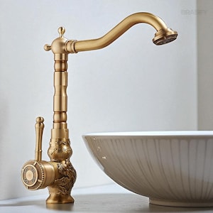 May include: A vintage-style brass faucet with an arched spout and decorative detailing. The faucet features a lever handle and a round dial with ornate patterns. The design includes floral and scrollwork elements, adding to its antique aesthetic. The faucet is positioned next to a white ceramic bowl.