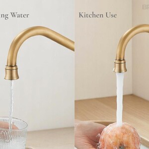 May include: A close-up of a gold-colored faucet with water flowing into a glass and onto a piece of fruit. The text "Drinking Water" and "Kitchen Use" are visible. The faucet has a curved spout and is set against a neutral background.