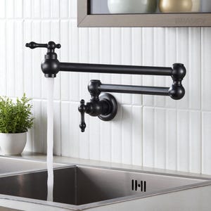 Puede incluir: Un grifo de llenado de ollas montado en la pared, de color negro mate, con un diseño de brazo oscilante. El grifo está situado sobre un fregadero de acero inoxidable, con agua que fluye del caño. El grifo está montado sobre azulejos blancos.