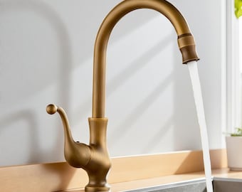 Puede incluir: Un grifo de cocina de color latón con un caño de arco alto. El agua fluye hacia un fregadero de acero inoxidable. El grifo tiene un diseño vintage con un caño curvo y un mango de palanca. La marca "BRASIFY" es visible en la esquina superior derecha.