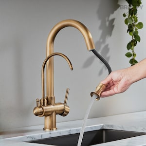 May include: A gold-toned kitchen faucet with a high arc spout and a pull-down sprayer. The faucet is mounted on a marble countertop above a black sink. A hand is holding the sprayer, and water is flowing.