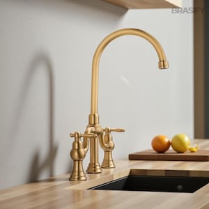 May include: A gold-colored kitchen faucet with a high arc spout and two lever handles. The faucet is installed on a light wood countertop. The image also shows a black sink, a cutting board, and two pieces of fruit.