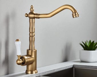 Puede incluir: Un grifo de cocina dorado con un caño curvo y un mango blanco. El grifo tiene un diseño clásico con detalles decorativos y está montado en una encimera. La marca "BRASIFY" es visible en la esquina superior derecha.