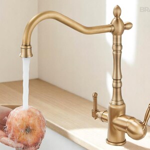 May include: A vintage-style brass kitchen faucet with a curved spout and ornate detailing. Water flows from the faucet onto a piece of fruit. The faucet has two handles and a decorative top. The brand name "BRASIFY" is visible.