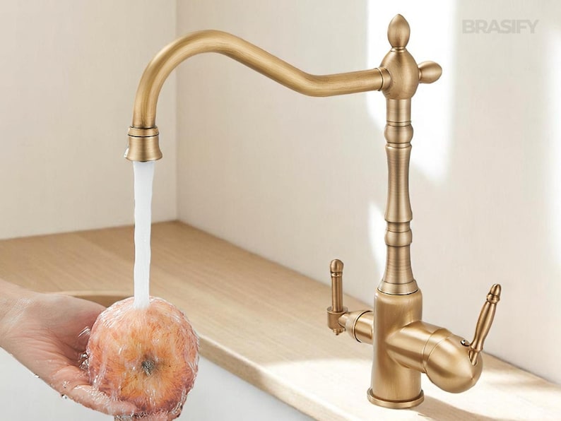 May include: A vintage-style brass kitchen faucet with a curved spout and ornate detailing. Water flows from the faucet onto a piece of fruit. The faucet has two handles and a decorative top. The brand name "BRASIFY" is visible.