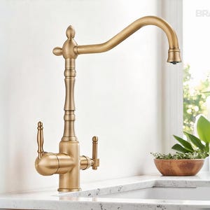 May include: A gold-toned kitchen faucet with a curved spout and dual handles. The faucet has an ornate design with decorative detailing. The brand name "BRASIFY" is visible in the upper right corner. The faucet is set against a white countertop.