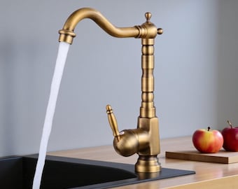 Puede incluir: Un grifo de cocina de latón antiguo con un caño curvo y una manija de palanca. El agua fluye del caño a un fregadero negro. El grifo tiene una columna decorativa y un diseño clásico. La palabra "BRASIFY" es visible en la esquina superior derecha.