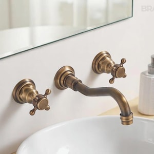 May include: Antique brass wall-mounted faucet set with cross handles. The faucet has a curved spout and is installed above a white ceramic sink. A white soap dispenser is visible to the right.