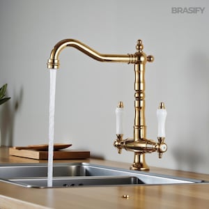 Puede incluir: Un grifo de cocina dorado con un caño curvo y manijas de porcelana blanca. El agua fluye del caño a un fregadero de acero inoxidable. La marca "BRASIFY" es visible en la esquina superior derecha.
