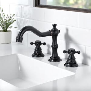 May include: A black bathroom tap with a curved spout and cross-head handles. The tap is set on a white countertop next to a white rectangular sink. A small potted plant is in the background.