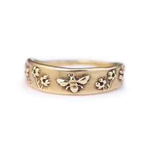 May include: A gold-colored ring with a bee and floral design. The ring features a detailed relief of a bee in the center, flanked by floral patterns on either side. The ring has a smooth, polished finish.