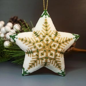 May include: A white, beaded star-shaped Christmas ornament with a gold and green snowflake design. The edges are trimmed in green beads, and a gold string is attached for hanging. The ornament is a festive holiday decoration.