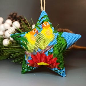 May include: A beaded star-shaped ornament with a nature-inspired design. The ornament features a red flower at the bottom, two yellow birds, and green leaves against a blue background. It is strung with a natural twine for hanging.