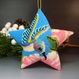 Peut inclure: Une étoile de Noël en perles bleues, roses et vertes avec un motif floral. L'étoile est faite de petites perles et a une ficelle dorée attachée au sommet.