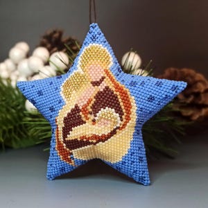 Może przedstawiać: Niebieska, gwiazdkowata ozdoba z koralikowym wizerunkiem Maryi i Dzieciątka Jezus. Ozdoba jest ozdobiona koralikami w kolorze złotym, brązowym i białym.