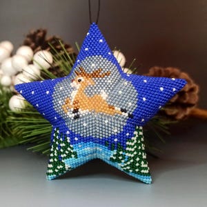 May include: A blue star-shaped Christmas ornament with a beaded reindeer design. The ornament features a white background with a blue border and a green pine tree design at the bottom.