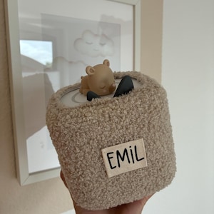 Peut inclure: Un récipient cylindrique en peluche beige avec une petite figurine d'ours endormi sur le dessus. Le récipient porte une étiquette en tissu avec le mot "EMIL" en lettres noires. Une illustration encadrée de nuages est en arrière-plan.