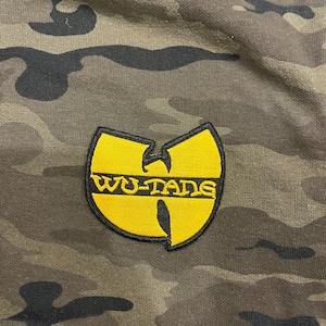 May include: Camouflage-patterned fabric with a yellow and black embroidered Wu-Tang Clan logo. The logo features the words "WU-TANG" in a bold, stylized font. The camouflage design includes shades of green, brown, and black.