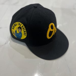 May include: Black baseball cap with a flat brim. The cap features a yellow embroidered "O" on the side and a circular patch with the text "BRING THE RUCKUS" and an image of a person in a blue outfit. The cap is made of a soft material.