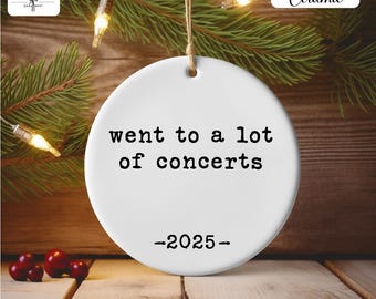 May include: White ceramic ornament with the text "went to a lot of concerts -2025-". The ornament is round and has a string for hanging. The background includes pine branches, string lights, and wooden planks.