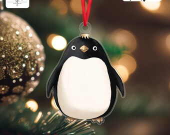 May include: A clear acrylic penguin Christmas ornament with a red ribbon for hanging. The penguin has a black head and wings, a white belly, and a gold beak. The word "Acrylic" is printed on the ornament.