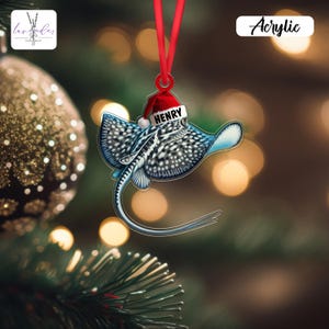 May include: A Christmas tree decoration made of acrylic, featuring a stingray design. The stingray is blue and white, wearing a Santa hat with the name "HENRY". It hangs from a red ribbon. The word "Acrylic" is visible.