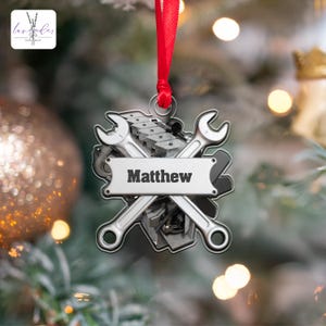Puede incluir: Adorno navideño plateado con un motor estilizado con llaves cruzadas y el nombre "Matthew" en una pancarta.