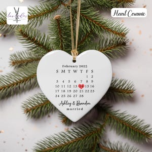 May include: White ceramic heart-shaped ornament with a calendar for February 2025. The 14th is highlighted with a red heart. The text "Ashley & Brandon married" is printed below the calendar.