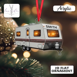 May include: A white and brown acrylic camper ornament with the name "SMITH" on the front. The ornament has a red ribbon for hanging and the text "2D FLAT ORNAMENT". The windows glow with a warm light, suggesting a cozy interior. The word "Acrylic" is also visible.