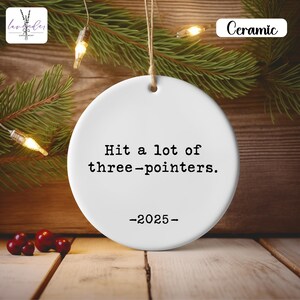 Puede incluir: Adorno navideño de cerámica blanca con el texto "Hit a lot of three-pointers. -2025-". El adorno está colgado de una cuerda contra un fondo de ramas de pino, luces y una superficie de madera. La palabra "Ceramic" está impresa en la parte superior derecha.