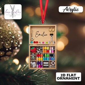 May include: A clear acrylic 2D flat ornament shaped like a sewing kit. The kit is open, showing spools of colourful thread, buttons, and sewing tools. The name "Emilia" is written on the wooden interior. A red ribbon is attached for hanging.