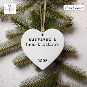 May include: A white heart-shaped ceramic ornament with the text "survived a heart attack -2025-". The ornament is hanging from a gold string against a backdrop of green pine branches. The words "Heart Ceramic" are in the upper right corner.