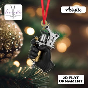 May include: Acrylic 2D flat ornament featuring a tattoo machine held by a black gloved hand. The machine is silver and gold, with the name "Michael" on the side. A red ribbon is attached for hanging.