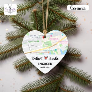 Puede incluir: Adorno de cerámica en forma de corazón con un diseño de mapa, con los nombres "Robert & Linda" y el texto "ENGAGED 06.18.2024" impresos. El adorno cuelga de una cuerda y se muestra sobre ramas de pino.