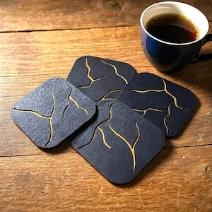 May include: Four square black coasters with gold-colored vein-like designs are arranged on a wooden surface. A blue coffee mug filled with dark liquid sits in the background, suggesting a coffee-themed product.