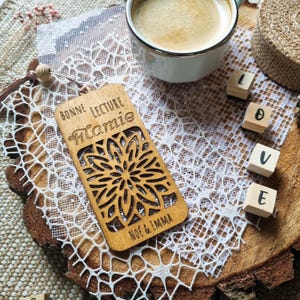 Peut inclure: Un marque-page en bois avec un motif floral découpé et le texte "Bonne Lecture Mamie" et "Noe & Imma". Le marque-page est posé sur une nappe en dentelle blanche, à côté d'une tasse de café et de cubes de lettres en bois épelant "LOVE".