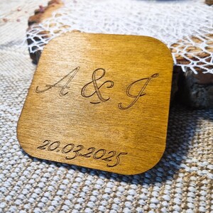 May include: A square wooden coaster with rounded corners. The coaster is engraved with the letters "A & J" and the date "20.03.2025" in a cursive font. The wood has a warm, light brown tone.