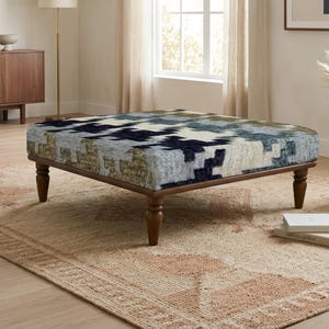 Pode incluir: Um otomano quadrado com um padrão geométrico pixelizado multicolorido em tons de azul-marinho, creme e verde-oliva suave. A peça é sustentada por quatro pernas de madeira torneada castanho-escuras que proporcionam uma base estável ao assento.