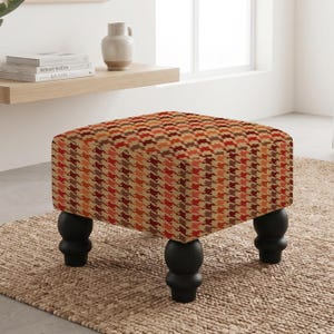 May include: A square ottoman featuring a multi-colored houndstooth pattern in shades of orange, red, and brown. The piece is supported by four black, rounded, bulbous wooden legs that provide a stable base for the upholstered seat cushion.