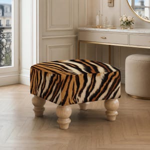 May include: A square ottoman featuring a bold tiger stripe pattern in shades of orange, black, and white. The piece is supported by four light-colored, rounded wooden legs with a turned design, providing a stable base for the upholstered seat.