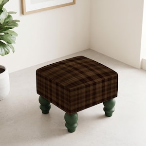 May include: A square ottoman featuring a dark brown and black plaid fabric upholstery. The piece is supported by four rounded, bulbous legs finished in a solid dark green colour, providing a stable base for the cushioned seat top.