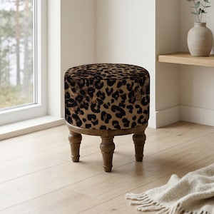 May include: A round ottoman with a leopard print top and wooden legs. The fabric features a tan background with black spots. The legs are a light brown color, and the ottoman is placed on a light wood floor.