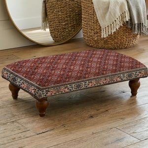 Può includere: Un poggiapiedi rettangolare caratterizzato da una superficie rosso scuro con un intricato motivo geometrico ripetuto. I lati sono decorati con un bordo ornamentale nei toni del crema, del blu e dell'arancione. Poggia su quattro gambe in legno tornito marrone scuro.