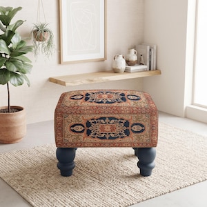 May include: A square ottoman featuring a traditional Persian-style rug pattern in shades of rust, navy blue, and beige. The piece is supported by four dark navy blue turned wooden legs with a rounded, bulbous shape at the base.