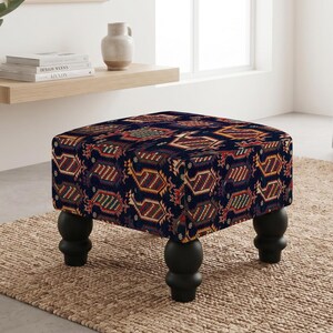 May include: A square ottoman featuring a dark navy blue fabric with a repeating geometric pattern in shades of red, yellow, and green. The piece is supported by four rounded, black wooden legs that provide a stable base for the upholstered seat.